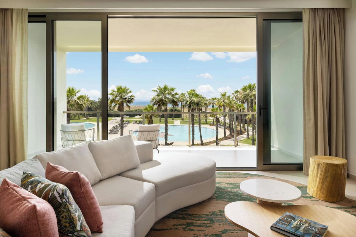 A modern living room with a curved white sofa, a round coffee table, and a view of a pool and palm trees through large sliding glass doors.
