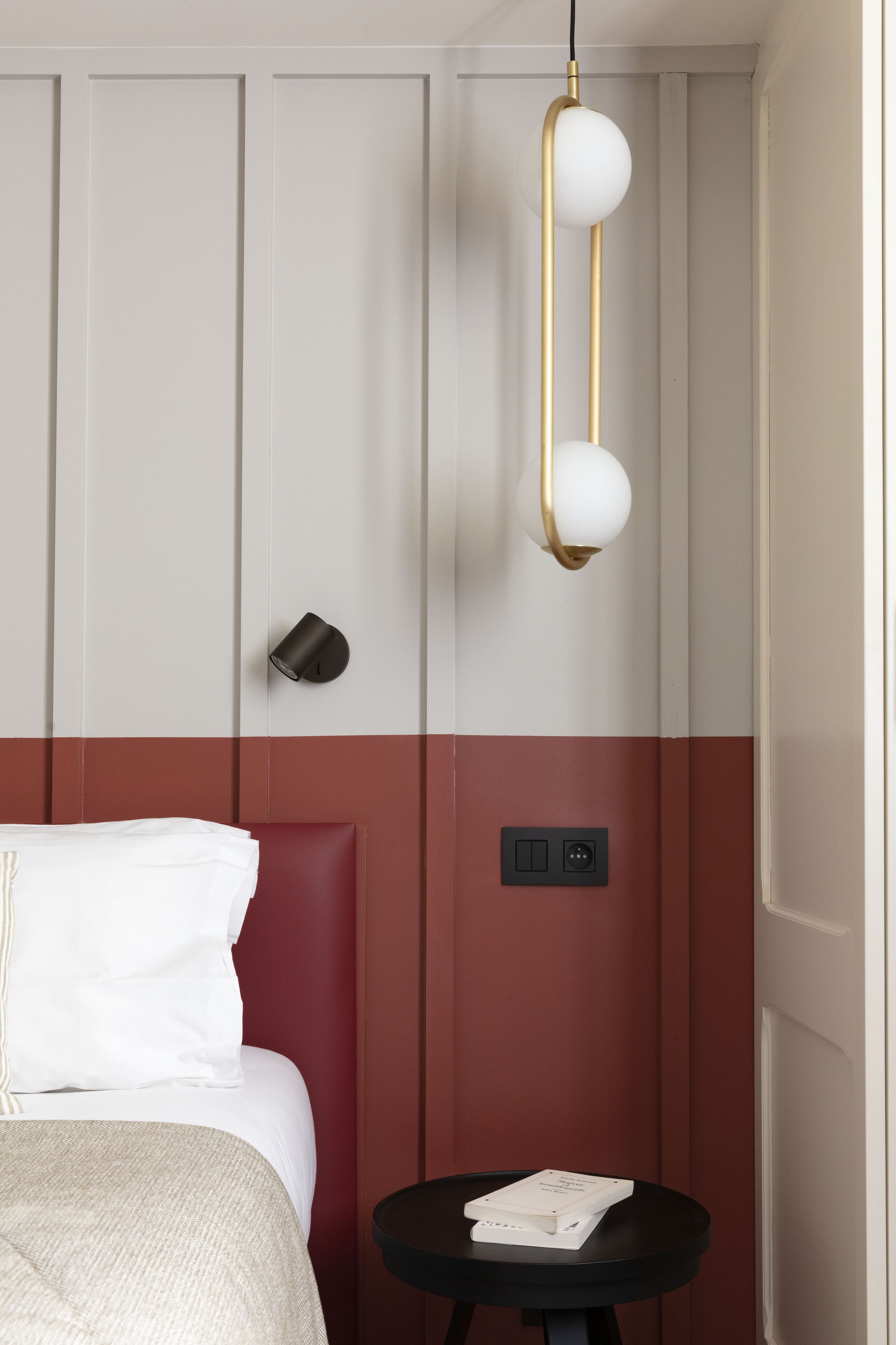 A hotel room with a bed, a small table with a book, a wall-mounted lamp, and a power outlet.