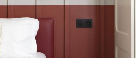 A hotel room with a bed, a small table with a book, a wall-mounted lamp, and a power outlet.