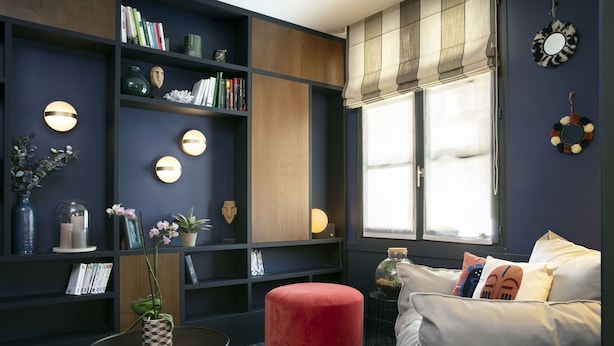 A modern living room with dark walls, a red ottoman, a white sofa, and a bookshelf filled with books and decorative items. - Hotel Bastille Spéria (Paris)