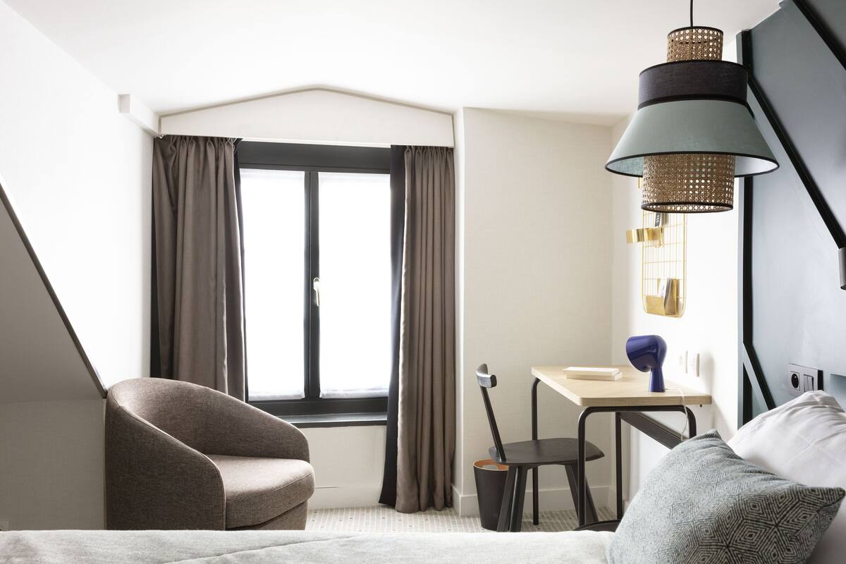 A modern hotel room with a curved sofa, a desk with a chair, a hanging lamp, and a large window with curtains.