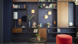 A modern living room with dark blue walls, wooden shelves, and a red ottoman. There are books, decorative items, and a small table with a vase of flowers. - Hotel Bastille Spéria (Paris)