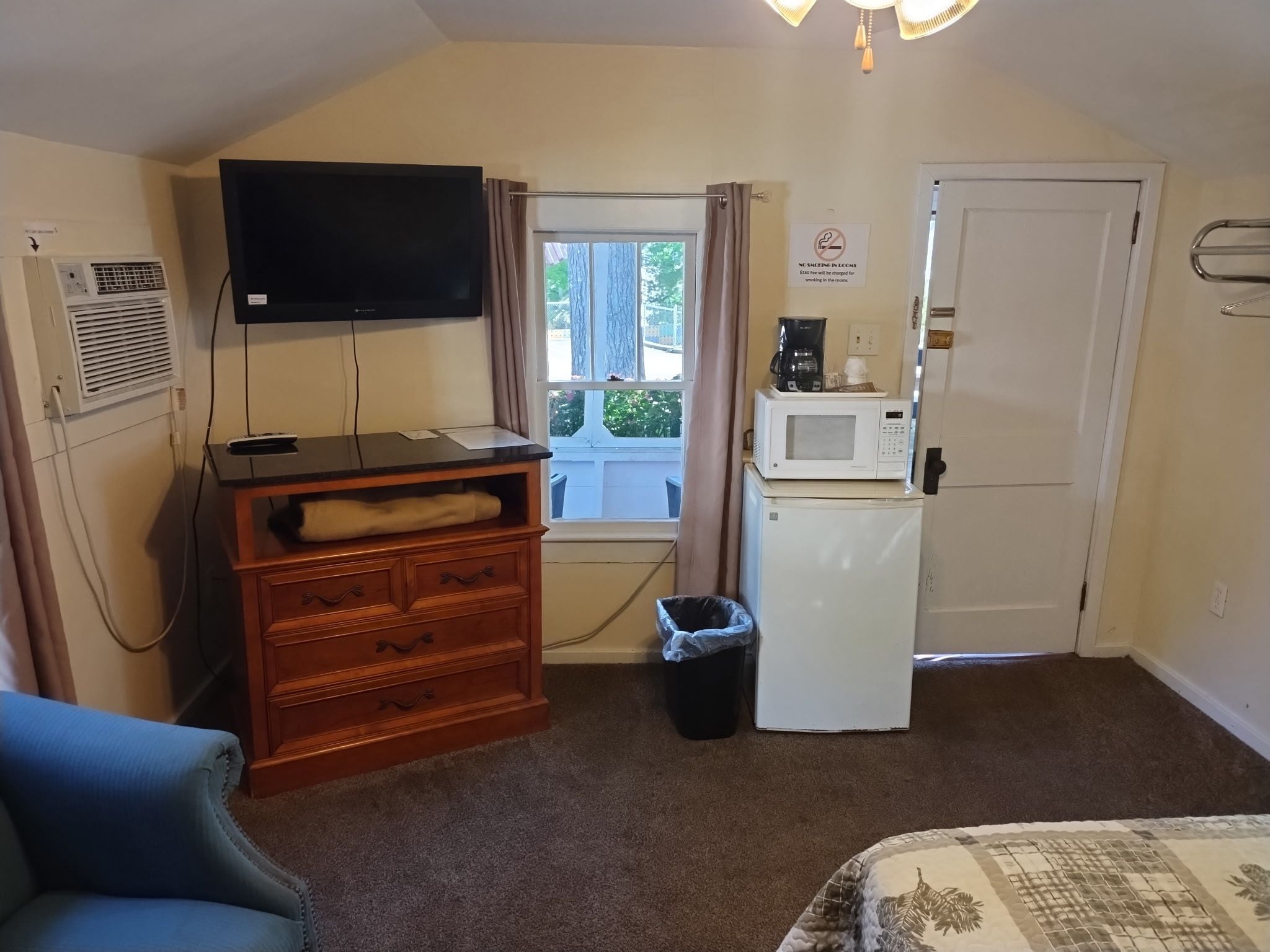 A small, furnished room with a flat-screen TV, a microwave, a refrigerator, a window with curtains, a trash bin, and a blue chair.