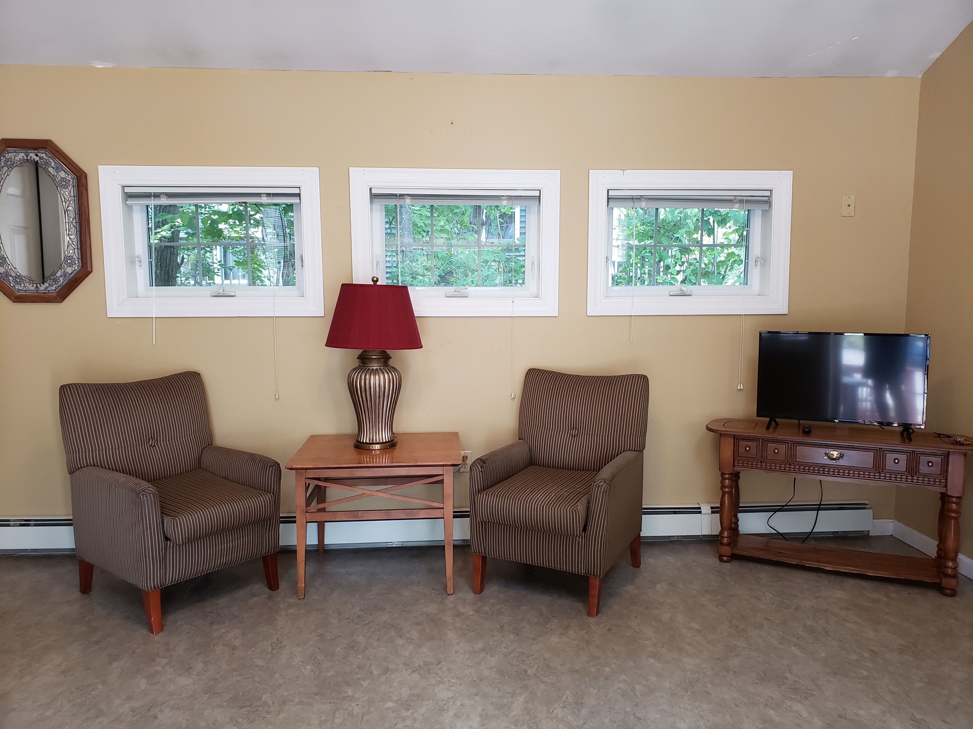 A room with two striped armchairs, a small wooden table with a red lamp, a television on a wooden stand, and three windows with white frames.