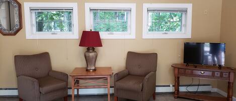 A room with two striped armchairs, a small wooden table with a red lamp, a television on a wooden stand, and three windows with white frames.