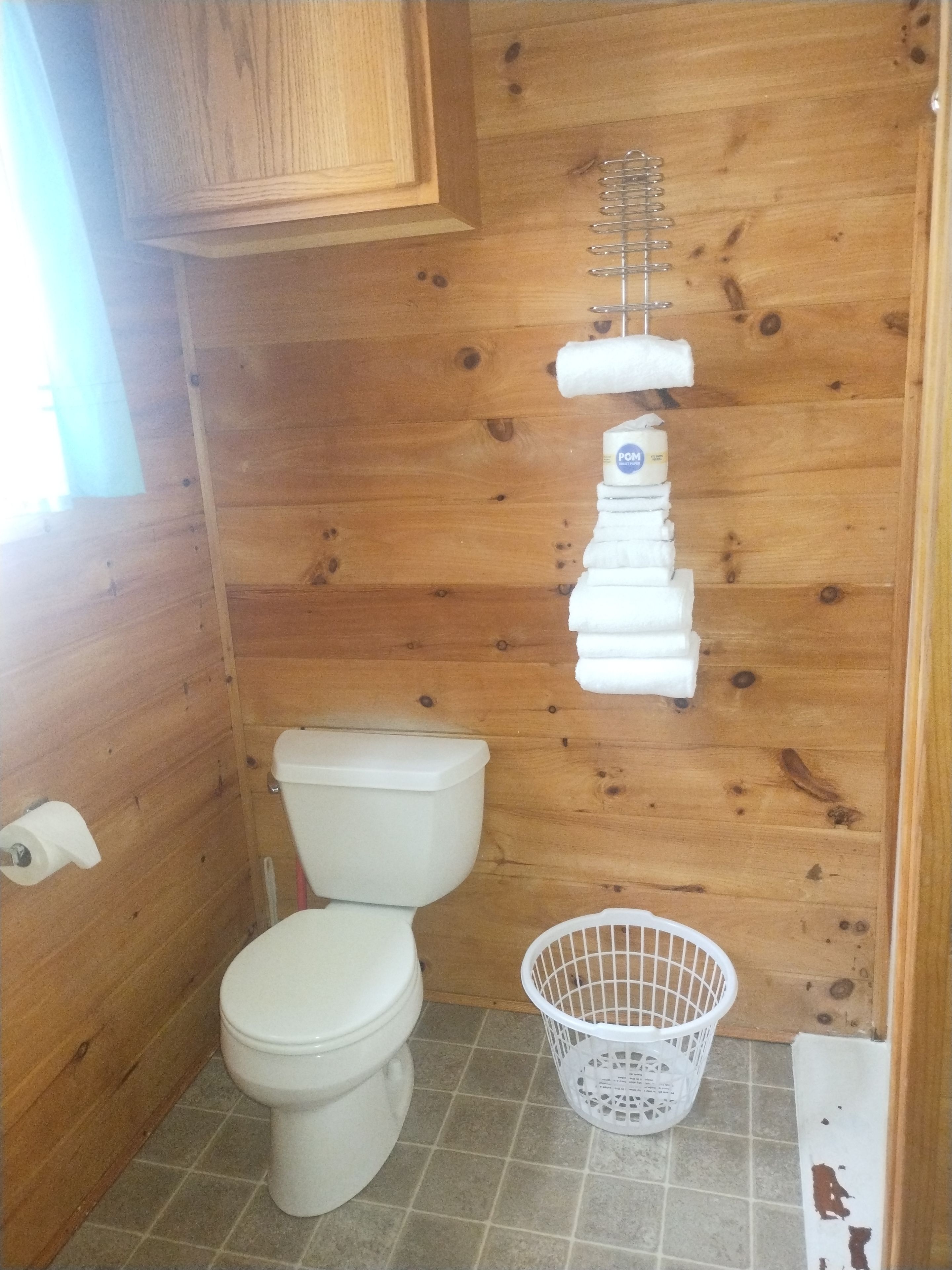 A small, wooden-paneled bathroom with a white toilet, a white toilet paper holder, and a white plastic laundry basket.