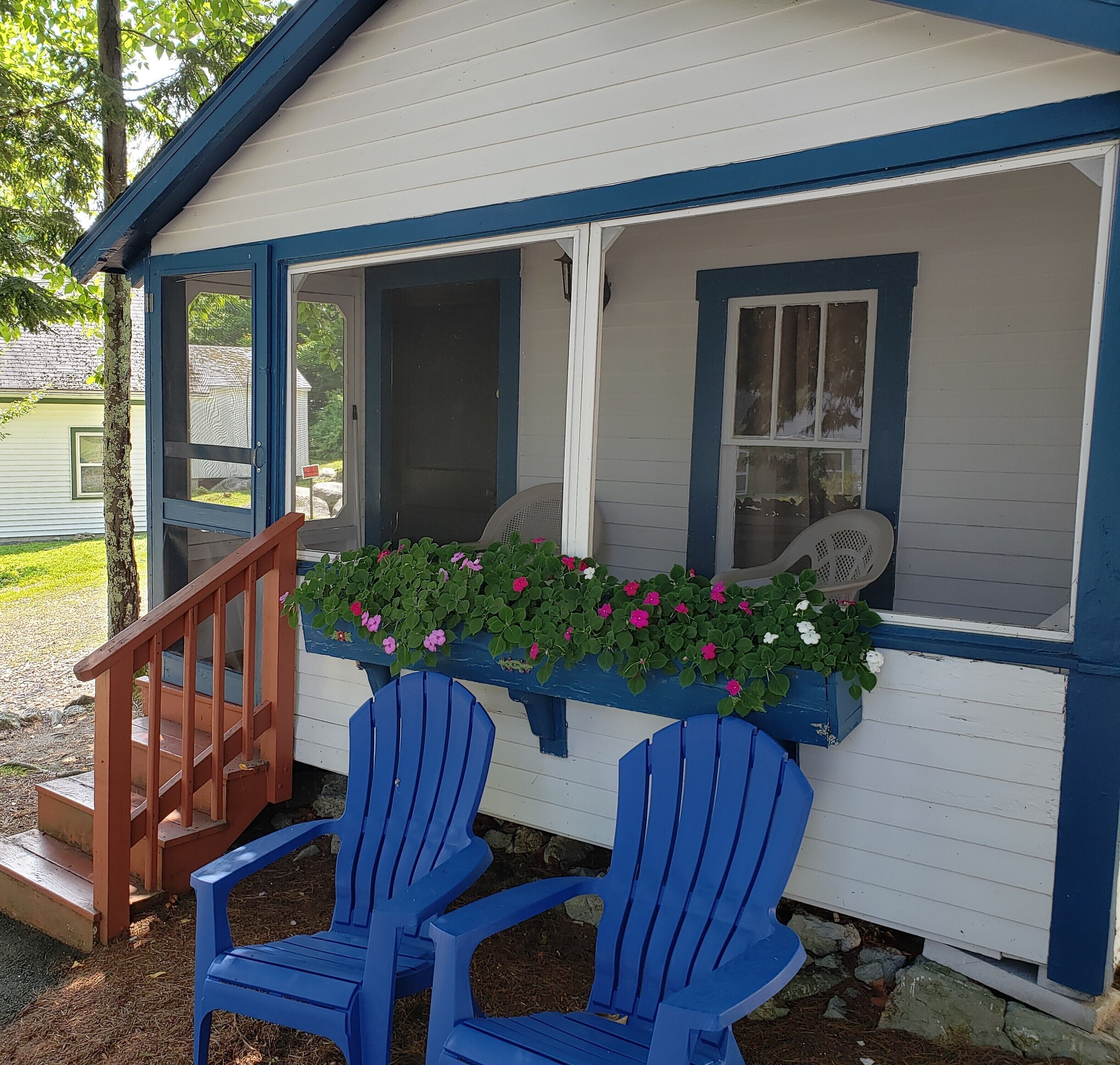 Uma casa pequena com varanda, duas cadeiras Adirondack azuis, uma escada de madeira e uma caixa de flores com flores cor-de-rosa.