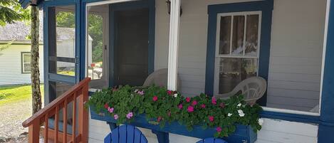 Uma casa pequena com varanda, duas cadeiras Adirondack azuis, uma escada de madeira e uma caixa de flores com flores cor-de-rosa.