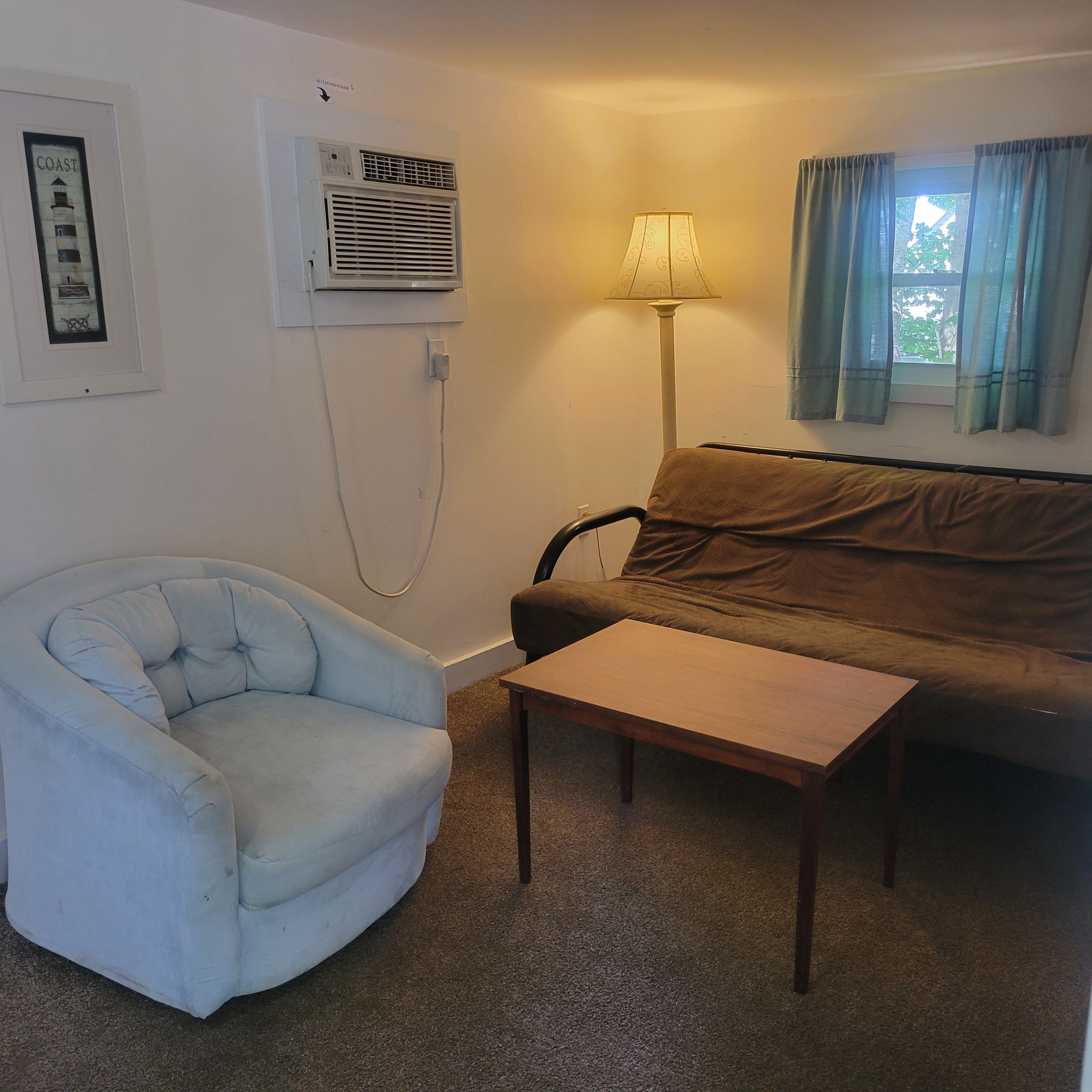 A room with a light blue armchair, a brown sofa, a wooden coffee table, an air conditioning unit, a lamp, and a window with curtains.