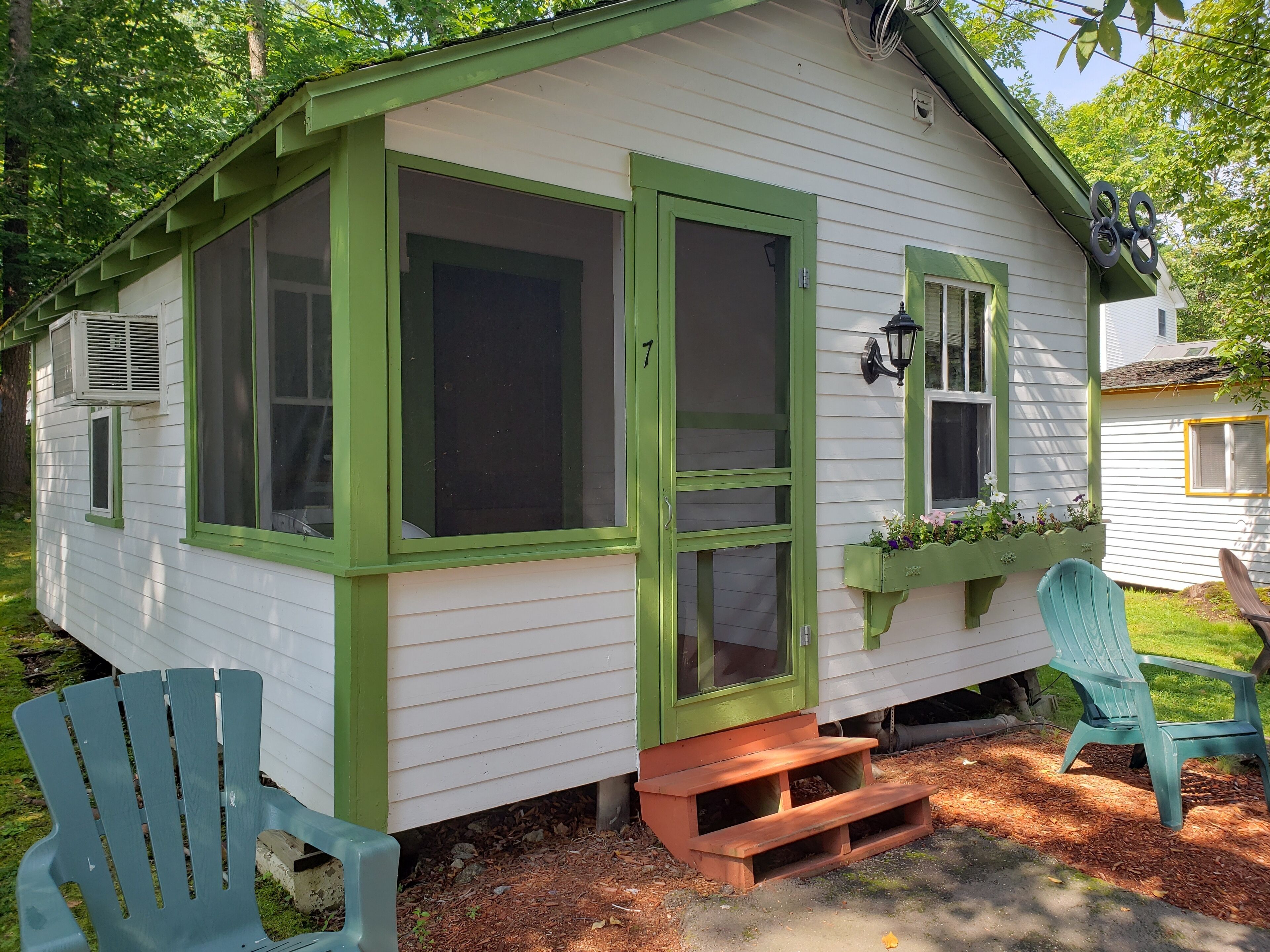 A small, single-story house with a green door and windows, a porch with steps, and a small garden area.