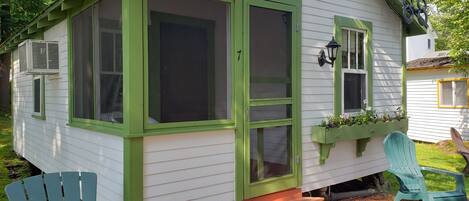 A small, single-story house with a green door and windows, a porch with steps, and a small garden area.