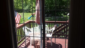 A view through a window with a metal grid, featuring a deck with white plastic chairs, a small round table, and a flag.