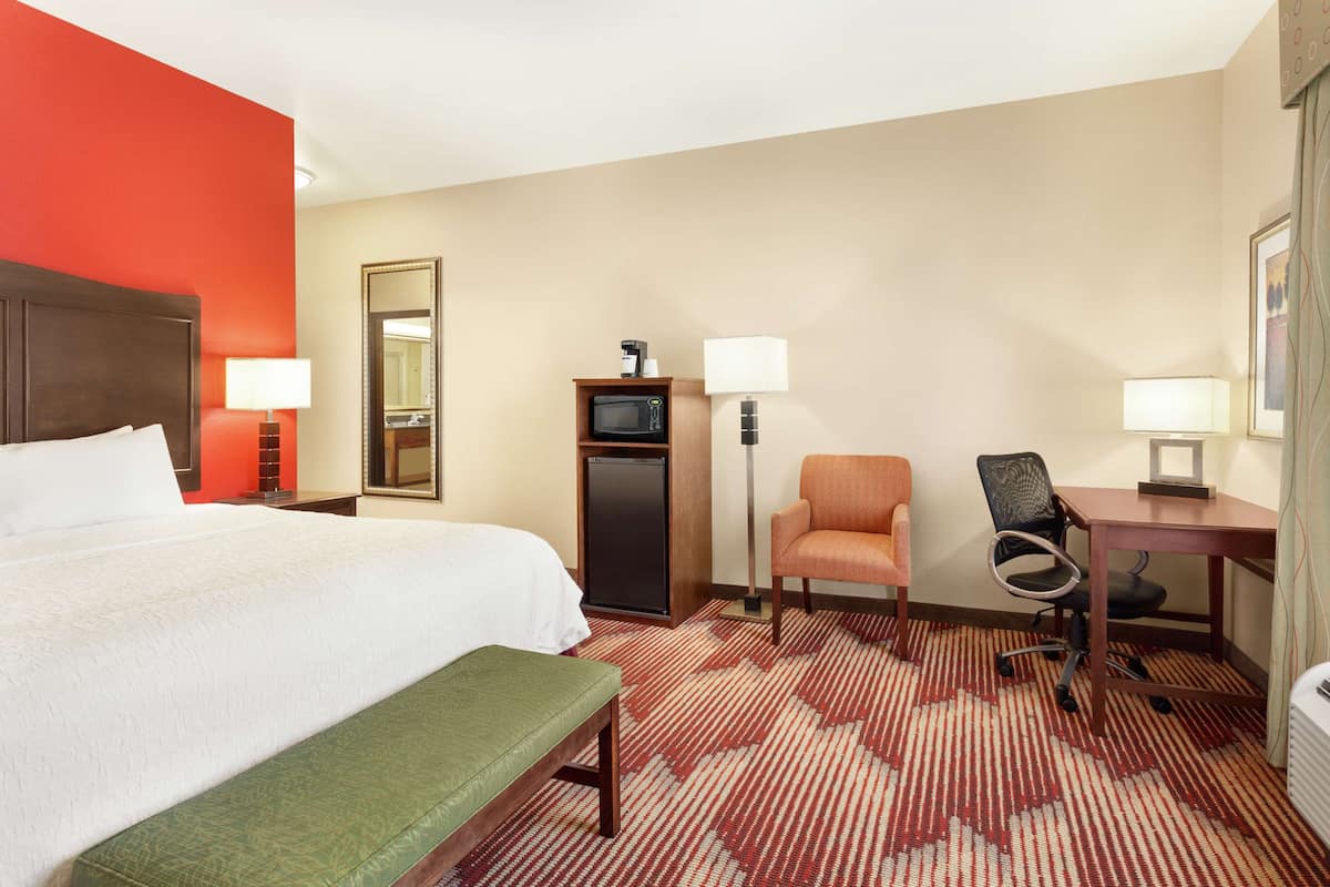 A hotel room with a bed, a desk, a chair, a bench, a microwave, a lamp, and a mirror.