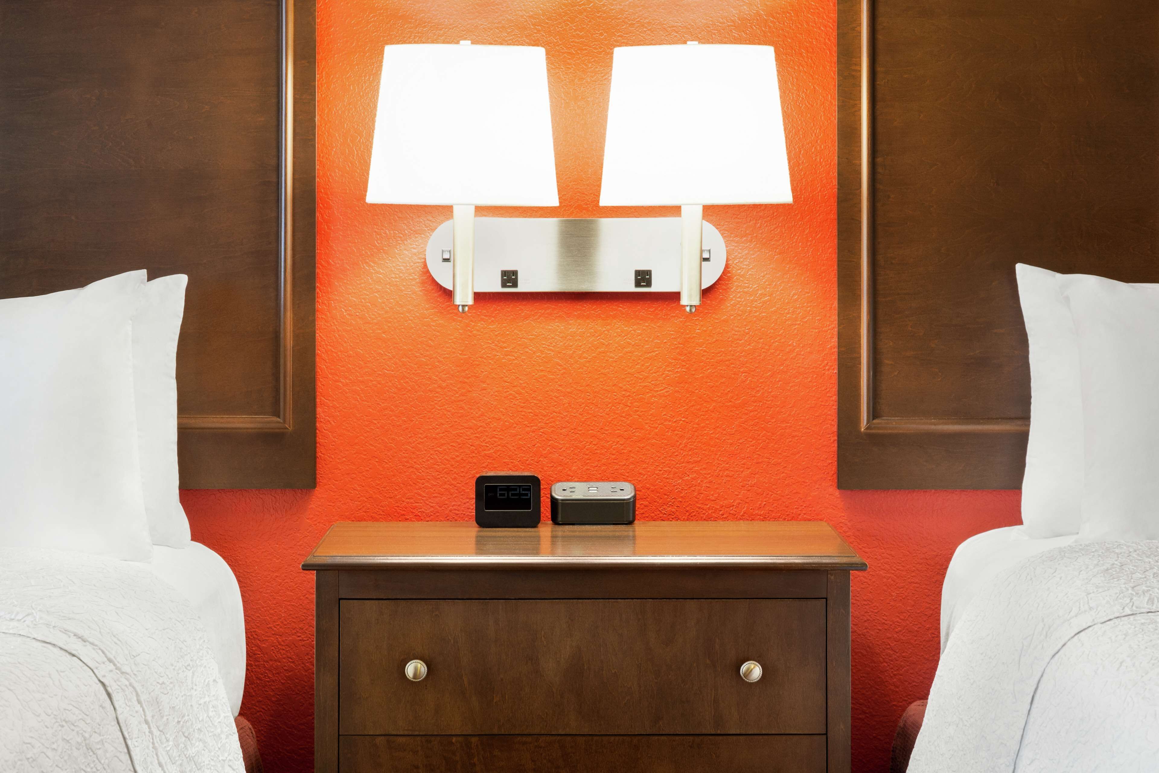 Una habitación de hotel con un mesita de noche de madera, dos mesitas auxiliares, un reloj digital y una lámpara de pared.