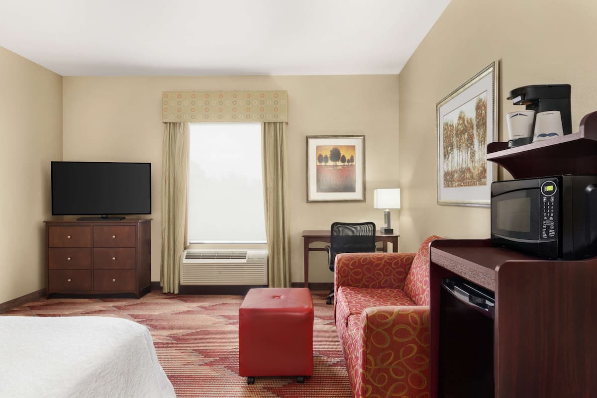 A hotel room with a bed, a red ottoman, a sofa, a television, a microwave, a coffee maker, a desk, a chair, a window with curtains, and a framed picture on the wall.