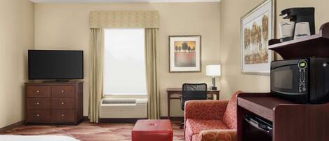 A hotel room with a bed, a red ottoman, a sofa, a television, a microwave, a coffee maker, a desk, a chair, a window with curtains, and a framed picture on the wall.