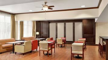A hotel lobby with a ceiling fan, multiple seating areas with tables and chairs, and large windows with curtains.