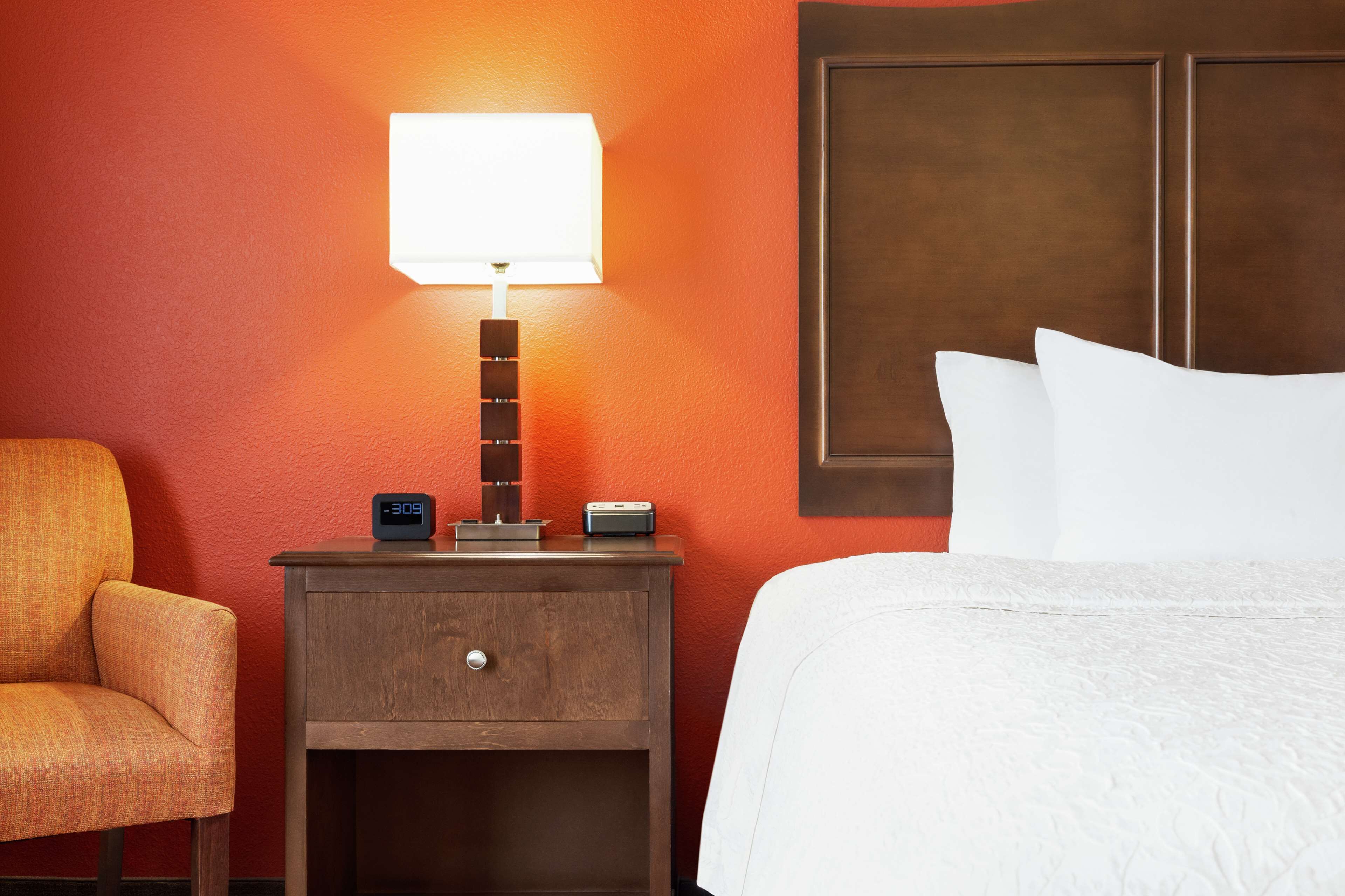A hotel room with a bed, a nightstand with a lamp, a clock, and a chair.