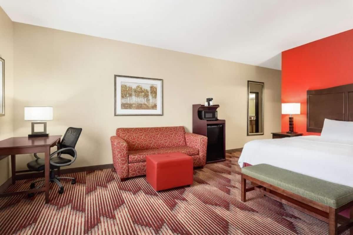 A hotel room with a bed, a red sofa, a small table with a chair, a desk, a mirror, and a picture on the wall.