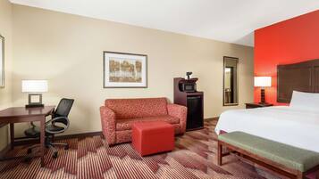 A hotel room with a bed, a red sofa, a desk with a chair, a small table, a microwave, and a framed picture on the wall.