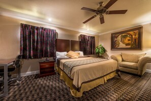 A hotel room with a bed, a desk, a chair, a sofa, a ceiling fan, and a painting on the wall.