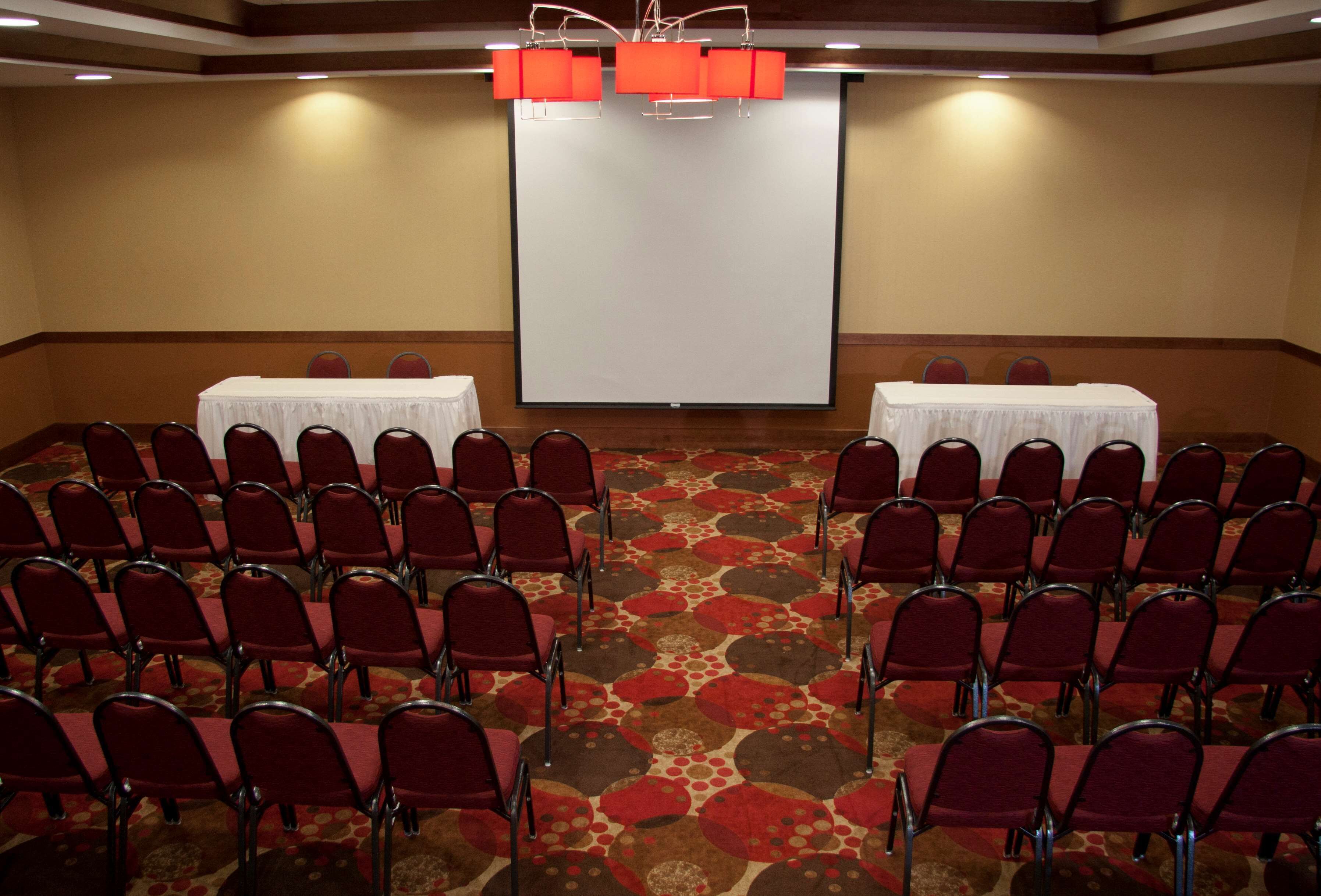 Una sala conferenze con file di sedie rosse, un grande schermo di proiezione e due tavoli con tovaglie bianche.