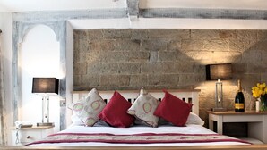 Une chambre avec un mur en pierre, un lit agrémenté d’oreillers rouges et à motifs, des lampes de chevet et une table de chevet sur laquelle se trouvent un téléphone et une bouteille.