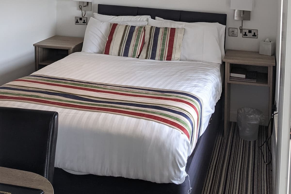 Habitación de hotel con una cama, dos mesitas de noche, una lámpara, un cesto de basura y un techo con vigas a la vista.