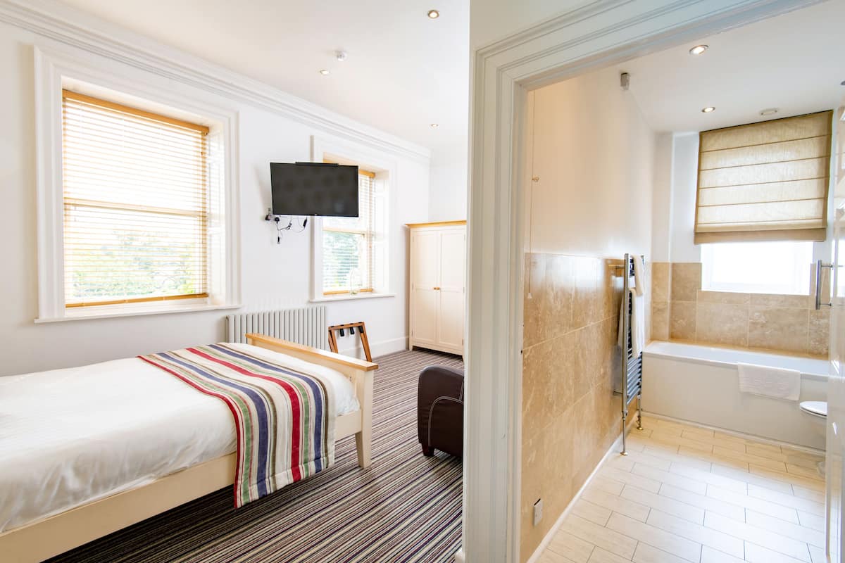 Habitación de hotel con cama, televisor, ventana con persianas, radiador, baño con bañera y ducha.