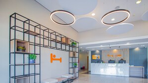 Un espace d’accueil d’hôtel au design moderne, comprenant un comptoir d’accueil, un coin salon avec un canapé et des fauteuils, ainsi qu’une étagère murale pour plantes.