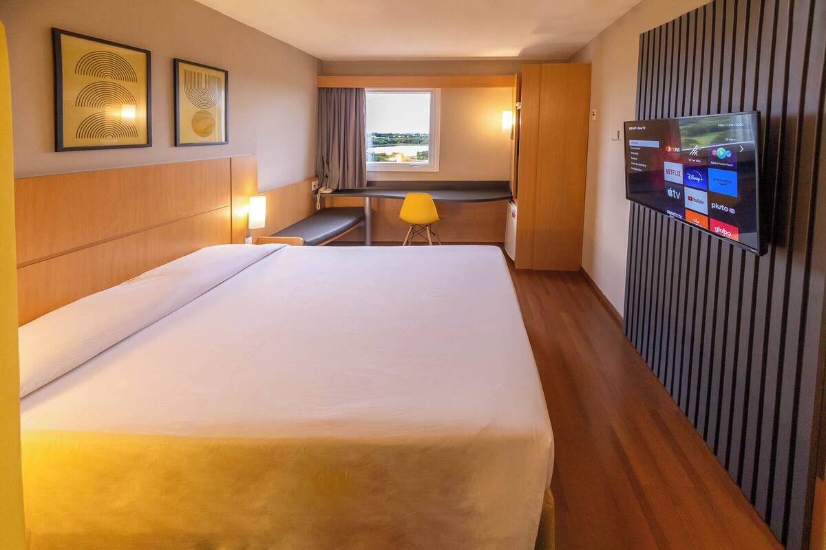 Une chambre d’hôtel équipée d’un lit, d’un bureau, d’une chaise, d’un téléviseur fixé au mur et d’une fenêtre avec des rideaux.