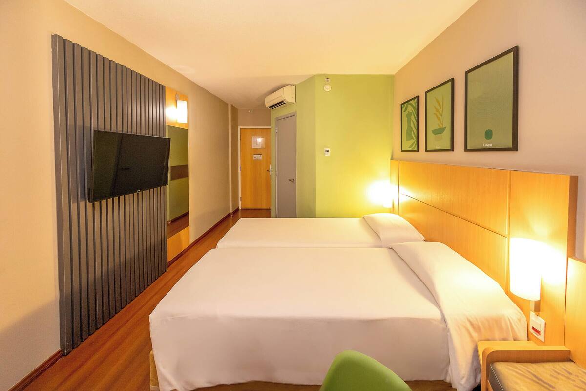 Une chambre d’hôtel avec deux lits, une télévision, une tête de lit en bois et des tableaux encadrés au mur.