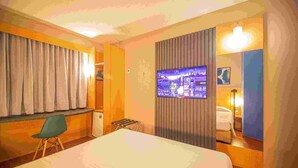 Une chambre d’hôtel comprenant un lit, une chaise, une petite table, une fenêtre avec des rideaux, un téléviseur fixé au mur et une table de chevet avec une lampe.