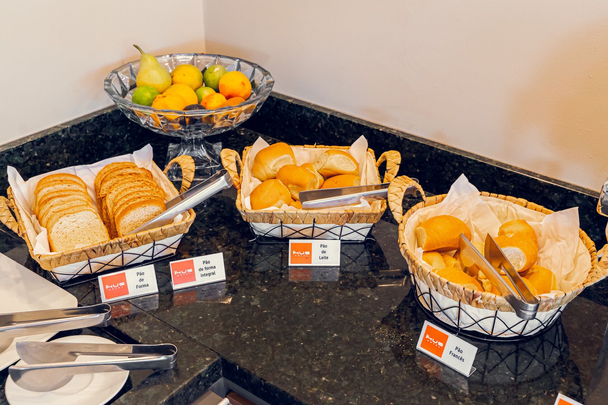A buffet with labeled bread baskets, a bowl of fruit, and serving utensils.