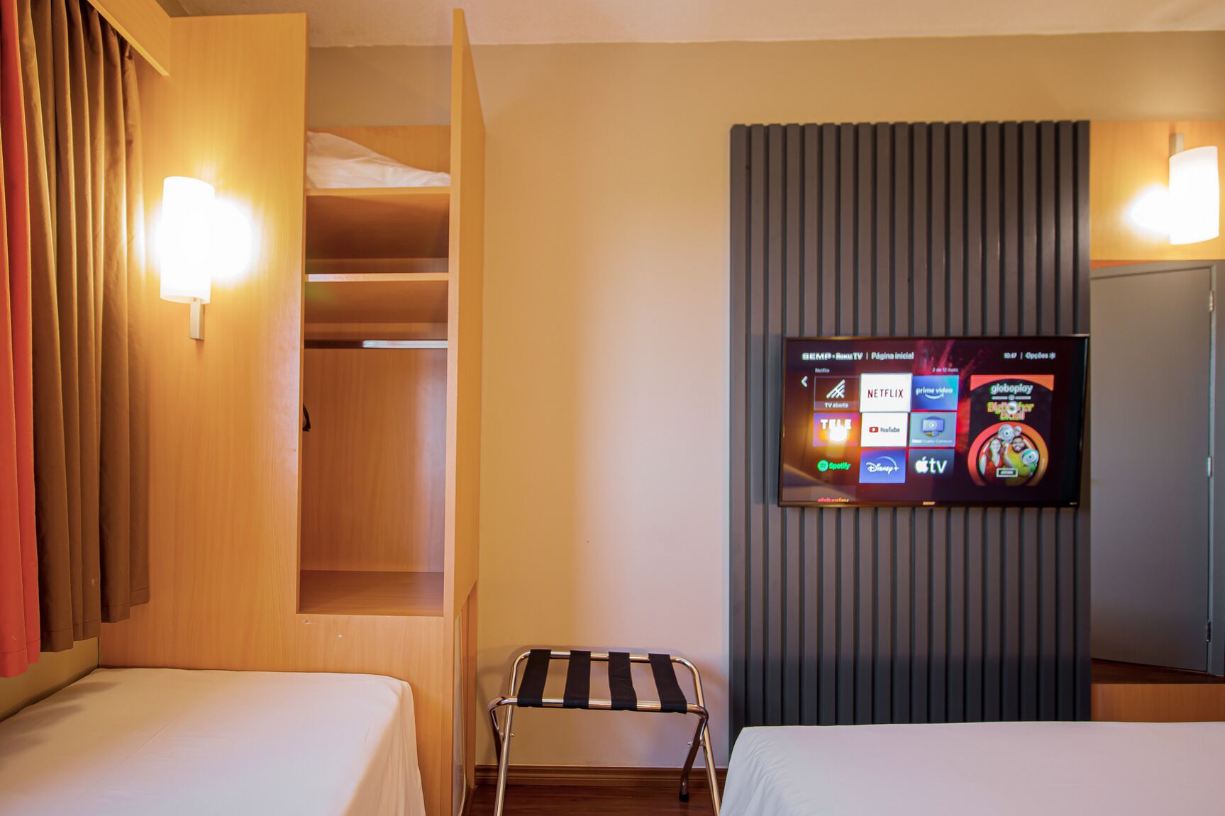 A hotel room with a TV displaying streaming services, a wardrobe, and a small seating area.