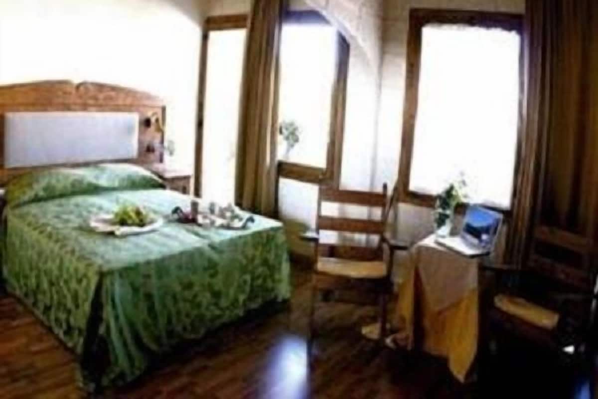 Quarto com cama, cadeira, uma pequena mesa com um laptop e janela com cortinas.