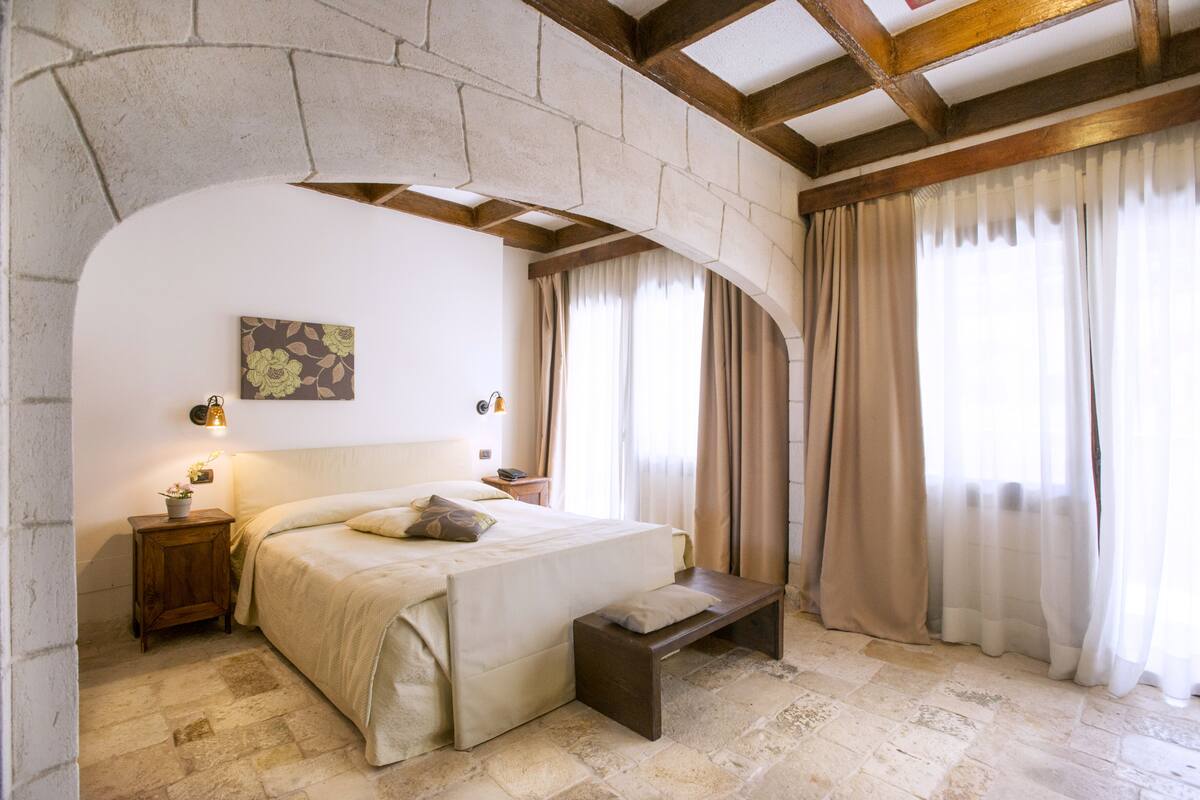 Um quarto com piso de pedra, uma cama, um banquinho, uma mesa de cabeceira e uma luminária fixada na parede.
