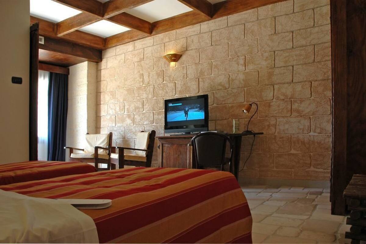 Quarto de hotel com duas camas, uma televisão sobre uma escrivaninha de madeira e uma parede de pedra.
