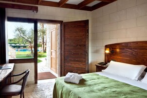 Habitación de hotel con cabecera de madera, cama con colcha verde, mesita de noche con lámpara y vistas a una zona de piscina al aire libre.