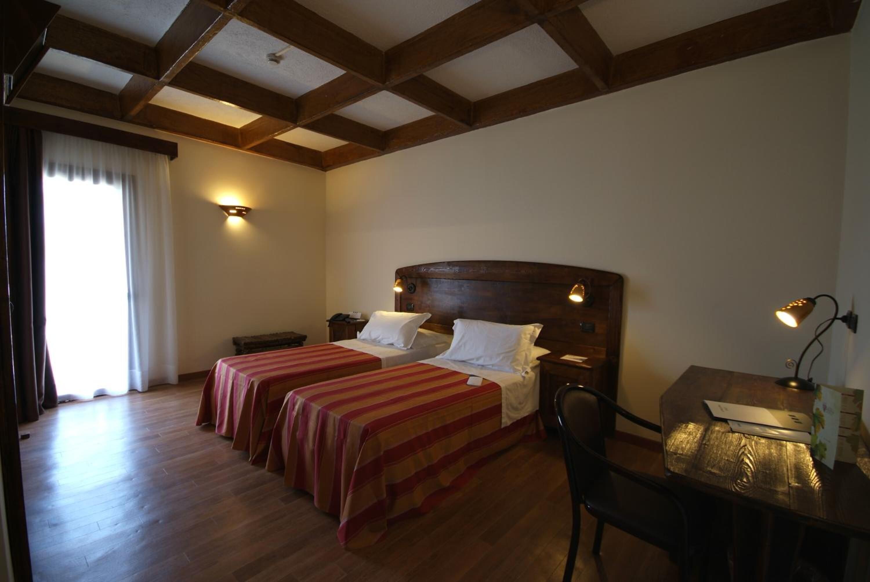 Une chambre d’hôtel avec deux lits, un plancher en bois, un bureau avec une lampe et une fenêtre avec des rideaux.