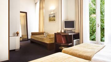 A hotel room with a bed, a sofa, a desk with a computer, a television, and a window with a view of greenery.