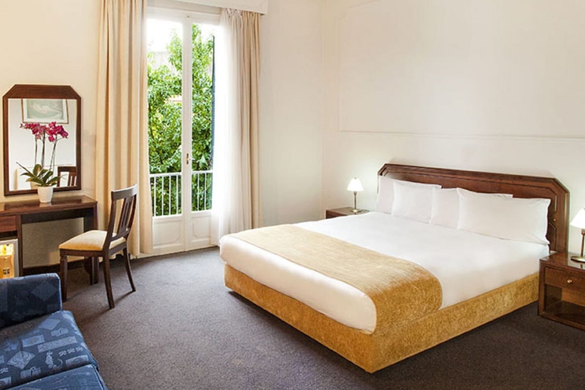 A hotel room with a bed, a desk with a computer, a chair, a sofa, and a window with a view of greenery.