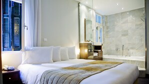 Une chambre d’hôtel avec un grand lit, une salle de bain avec baignoire et une fenêtre avec volets.