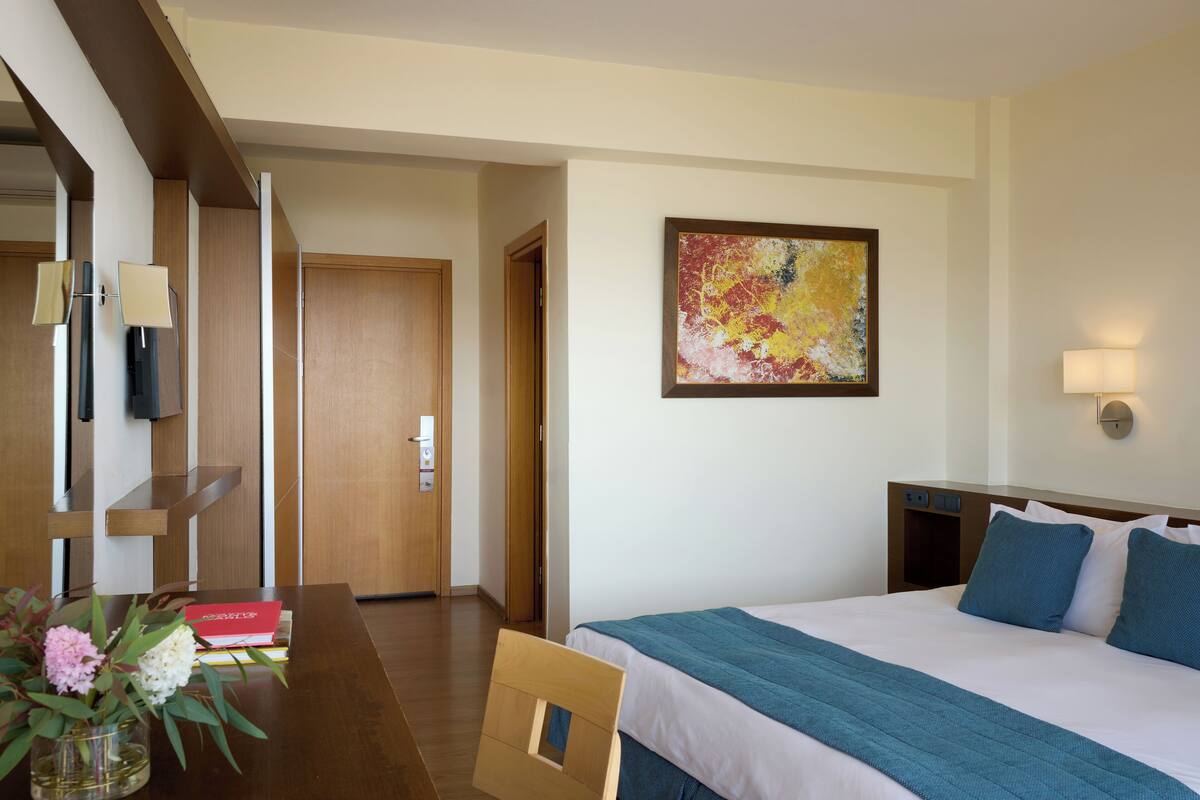 A hotel room with a bed, a desk with a vase of flowers, a painting on the wall, and a door leading to another room.