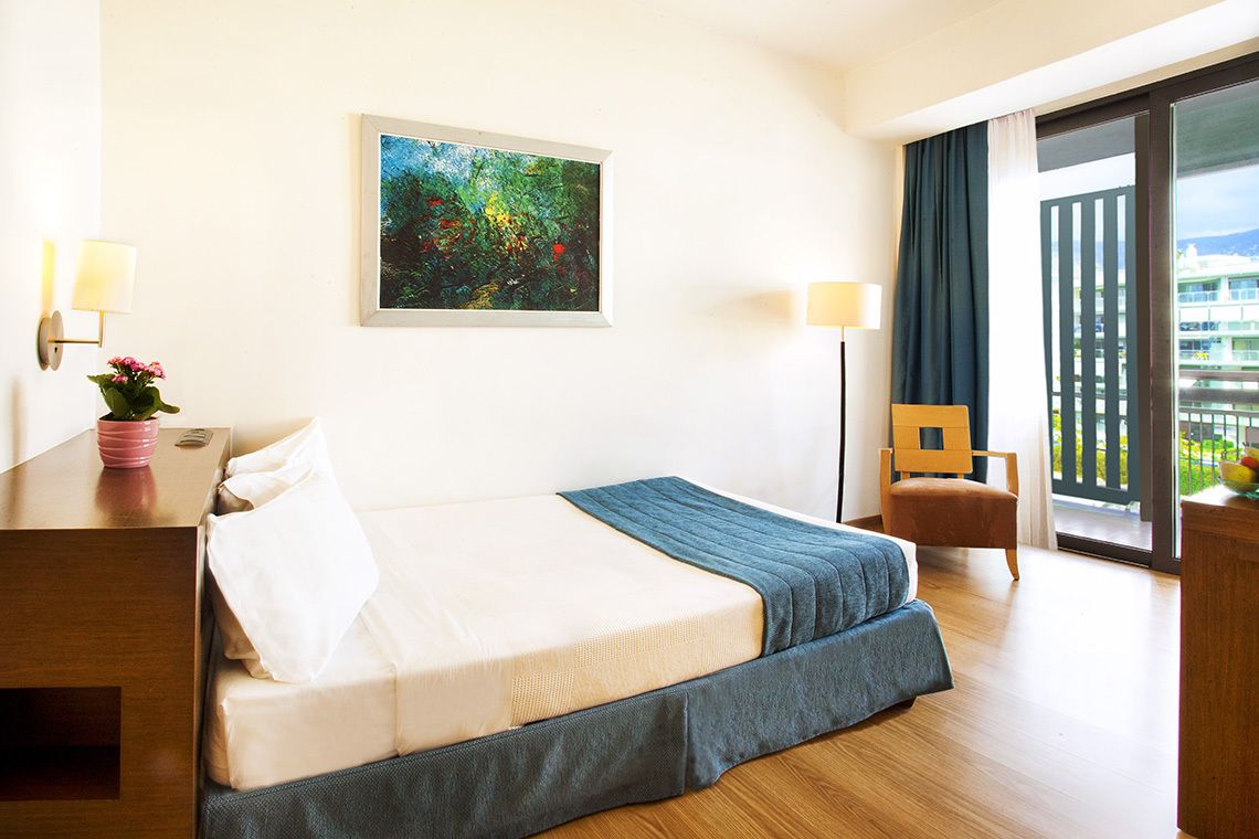 Une chambre d’hôtel avec un lit, un bureau doté d’une lampe, une chaise, un tableau au mur et un balcon avec vue.
