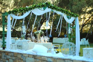 An outdoor wedding setup with white tables, a floral arch, and a stone wall.