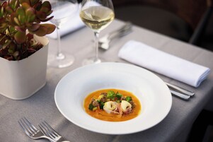 Un plato blanco con un plato de sopa, una copa de vino blanco y una pequeña planta en maceta sobre una mesa.