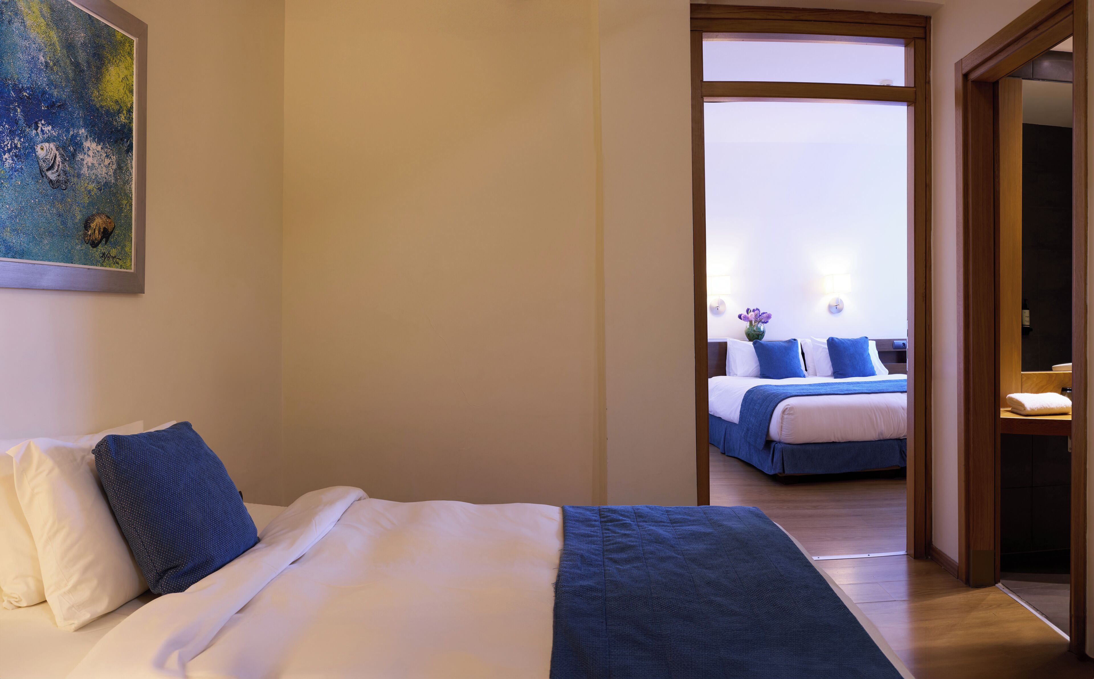 A hotel room with a bed, a painting on the wall, and a view into another room with a bed and a vase of flowers.