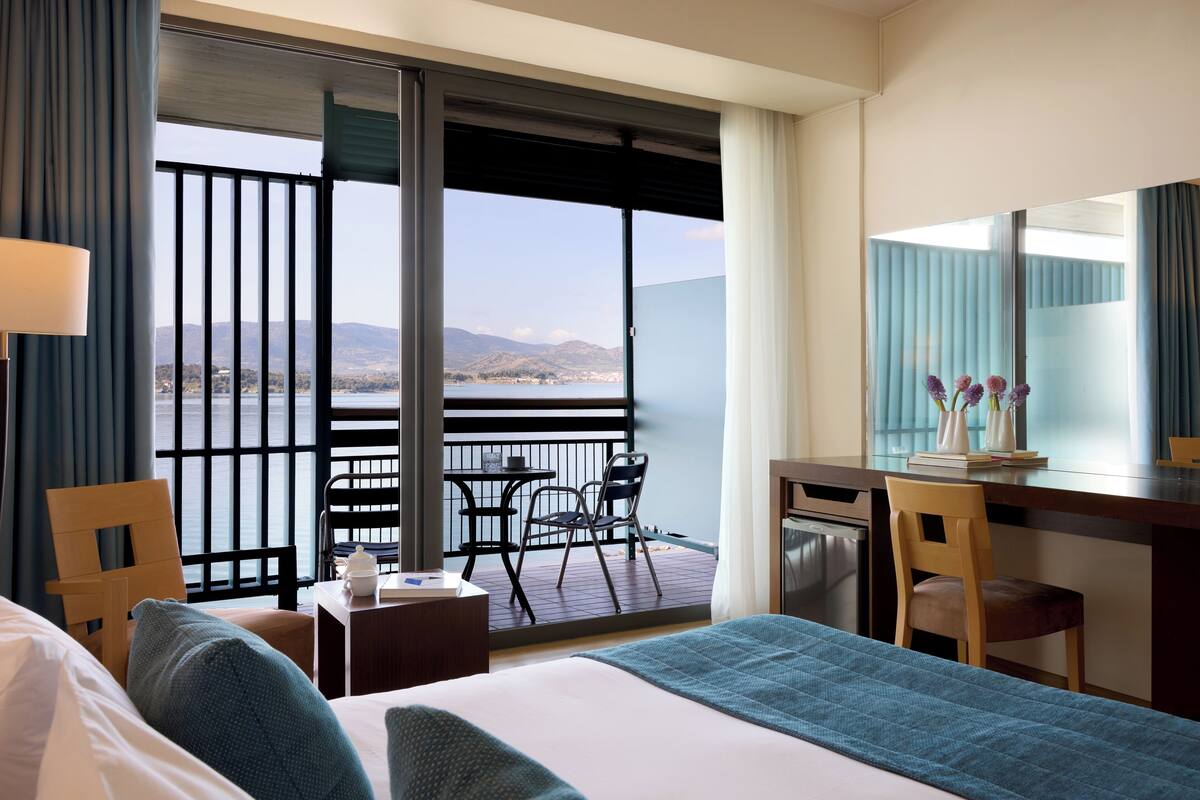 A hotel room with a bed, a desk, a chair, a small table, and a view of the sea.