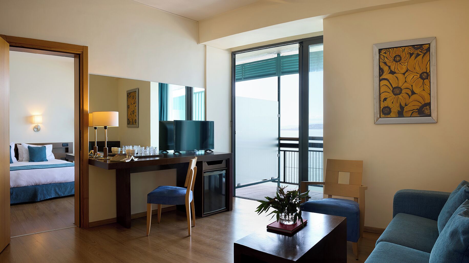Habitación de hotel con cama, escritorio con televisor, balcón con vista y un cuadro en la pared.